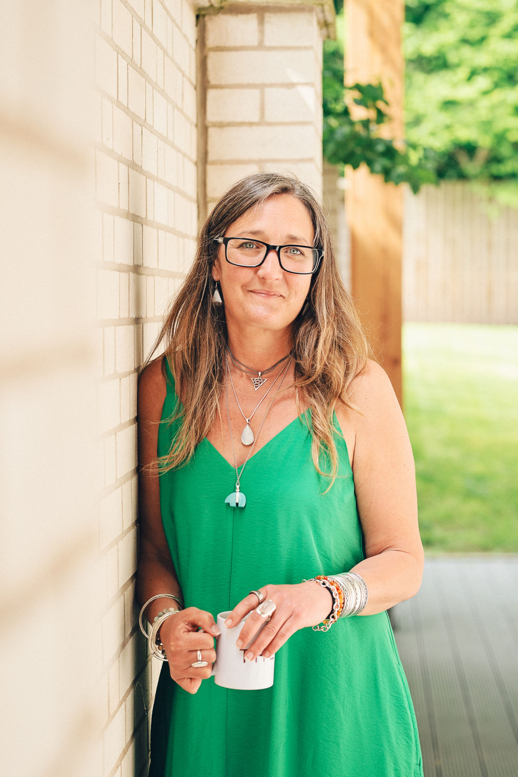 Ali Knight leaning against a white brick wall outdoors, holding a mug with both hands, wearing a green sundress, layered silver necklaces and multiple bracelets, smiling directly at the camera with a warm, relaxed expression.