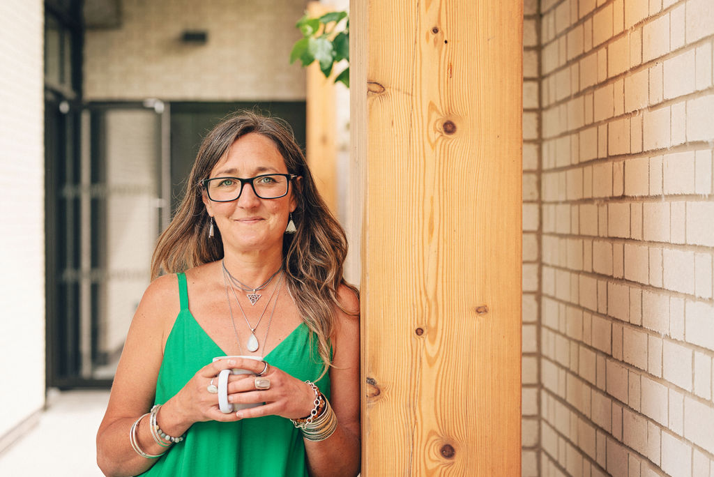 Ali Knight leaning against a wooden pillar next to a white brick wall, outdoors, holding a mug with both hands, wearing a green sundress, layered silver necklaces and multiple bracelets, smiling directly at the camera with a warm, relaxed expression.
