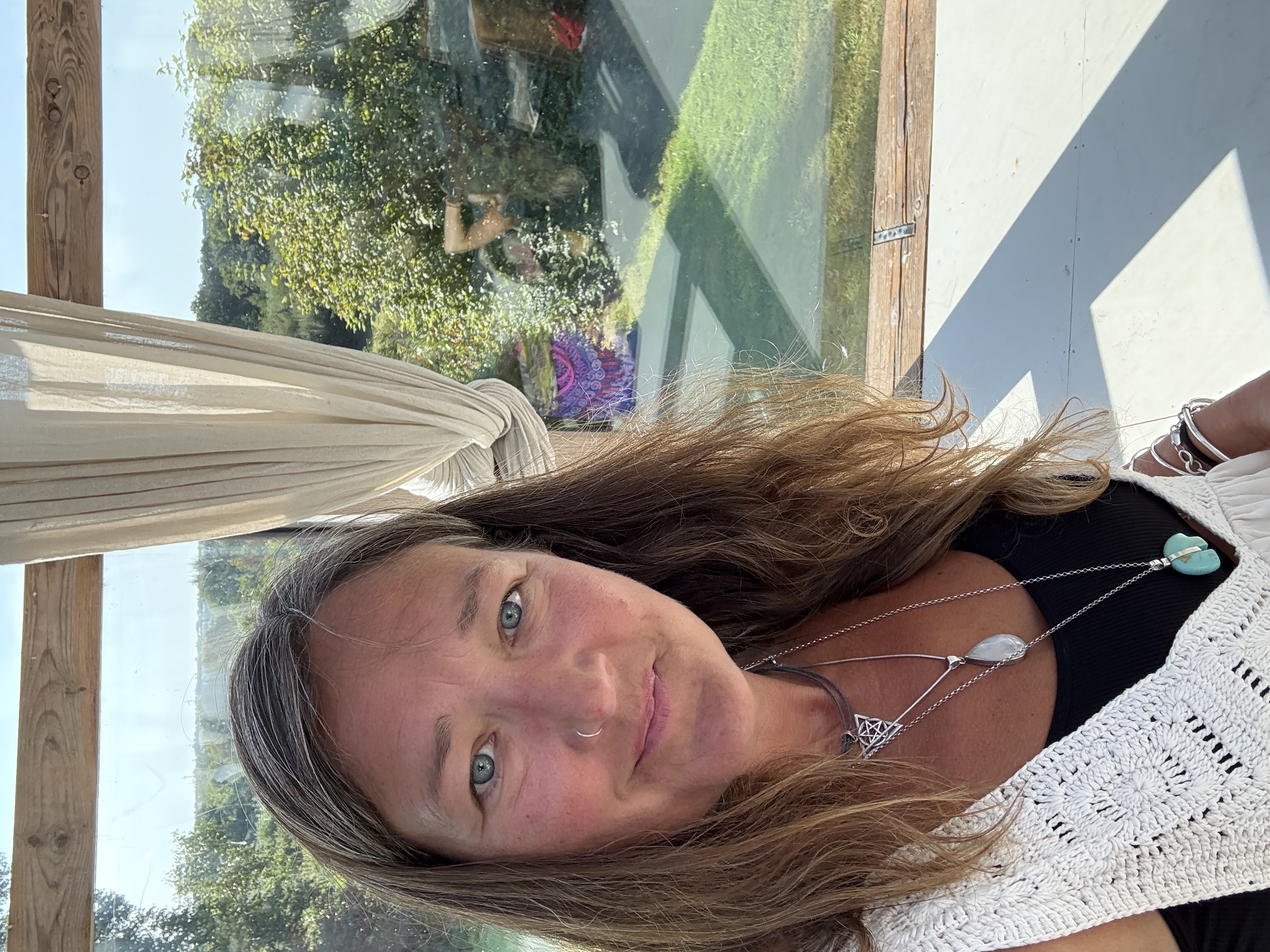 Ali Knight, a white woman with long hair and a nose ring, looking directly at the camera, wearing a white crochet top and layered silver necklaces including a turquoise pendant, with a large window behind her looking out onto a sunny garden where people can be seen gathered around a colourful mandala cloth.