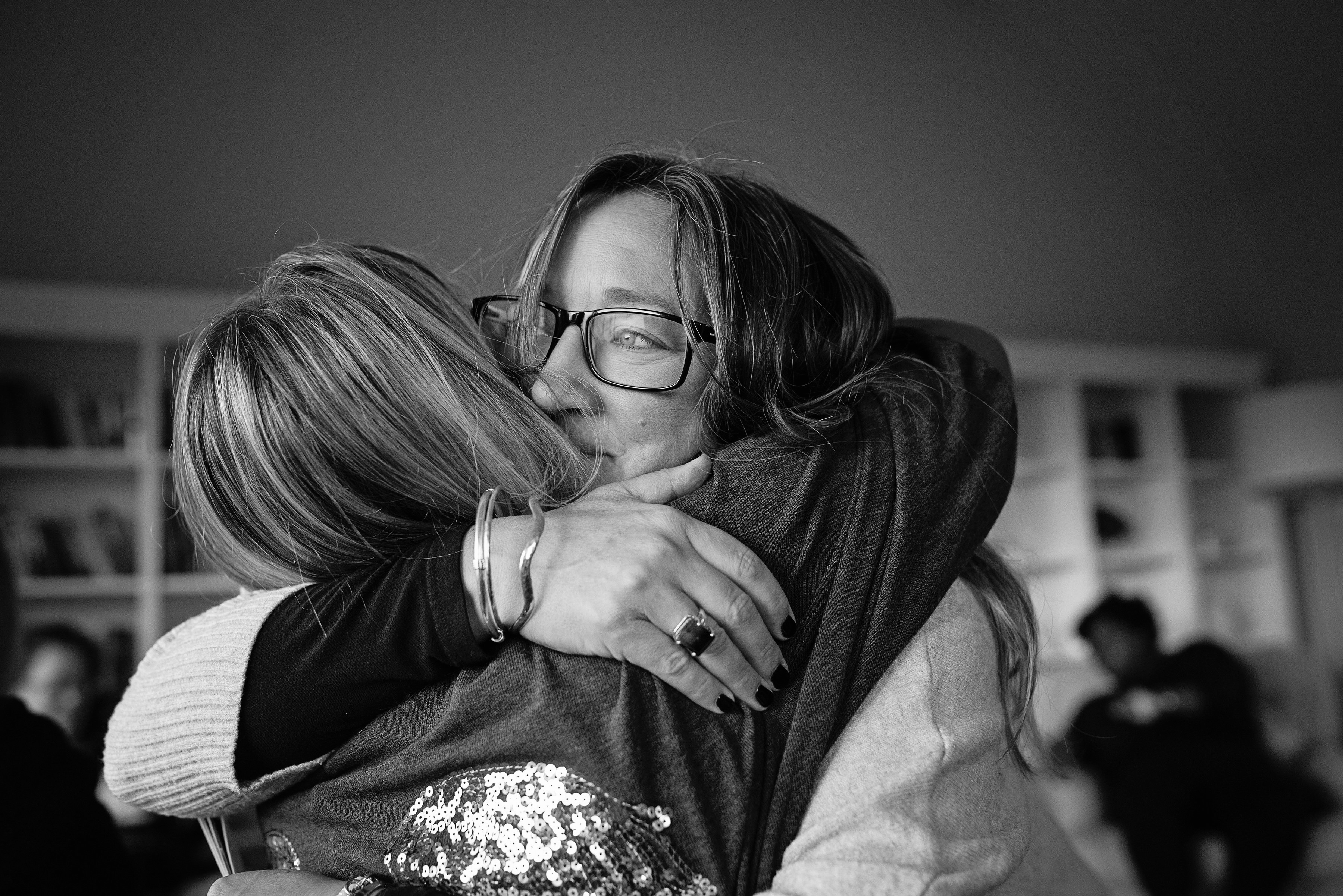 Ali Knight, a white woman with glasses, sharing a close, heartfelt embrace with another white woman in a softly lit indoor space, photographed in black and white, with other people visible in the background
