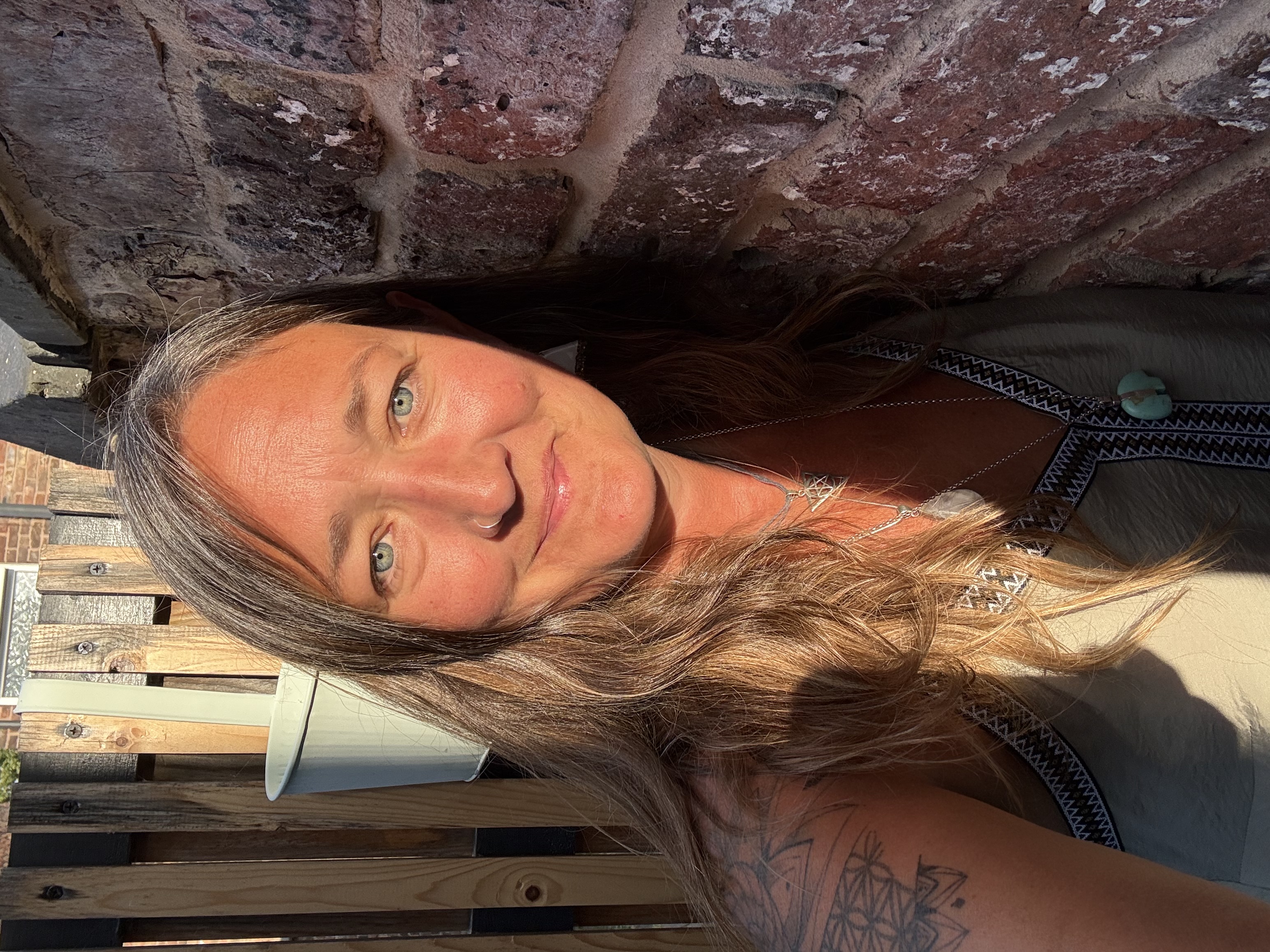 Ali Knight, a white woman with long blonde and grey hair and a nose ring, looking directly at the camera with a relaxed, warm expression, leaning against a red brick wall in golden evening light, wearing a patterned vest top and layered silver necklaces including a turquoise pendant, with a geometric tattoo visible on her upper arm