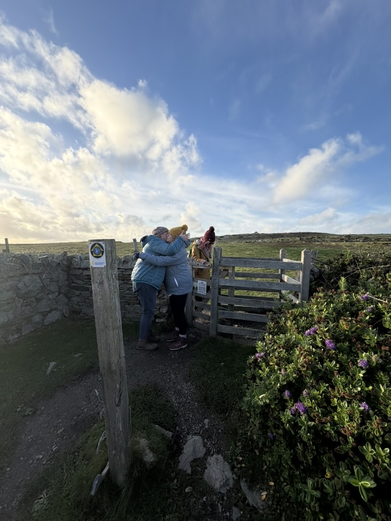Two white people embracing warmly at a wooden gate on a rugged moorland path during an Ali Knight retreat, wearing winter coats and woolly hats, with a wide open sky, dry stone walls and flowering heather visible around them