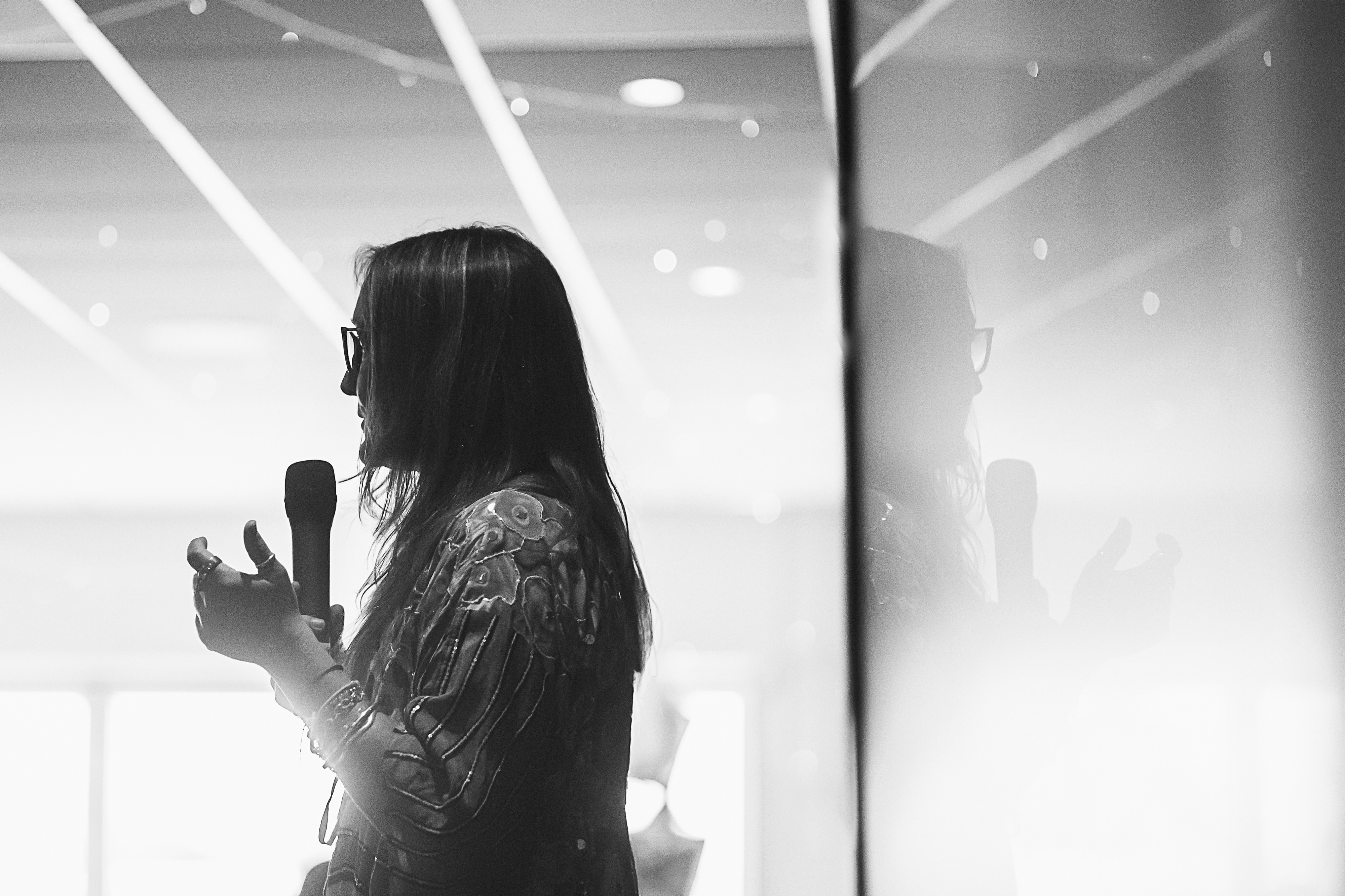 Ali Knight, a white woman with long hair and glasses, photographed in black and white from the side, speaking into a microphone with one hand raised expressively, wearing a patterned top and bracelets, with her reflection visible in a glass panel behind her