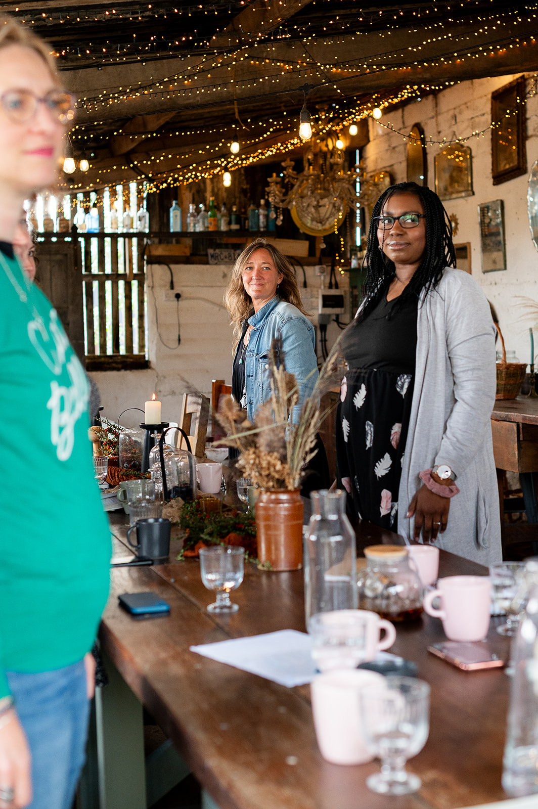 Ali Knight, a white woman with long hair, standing at a long wooden table during a group event in a warmly lit rustic space decorated with fairy lights, alongside a Black woman with braids and glasses, with candles, dried flowers, mugs and glassware on the table and a white woman in a green top partially visible in the foreground