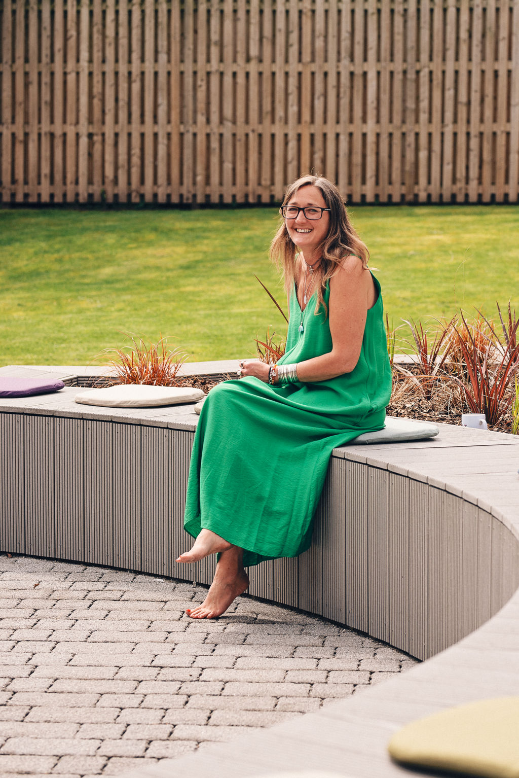 Ali Knight, a white woman with long blonde-flecked hair and glasses, sitting barefoot on a curved outdoor bench in a bright green maxi dress and silver jewellery, laughing and looking directly at the camera, with a lawn and ornamental grasses visible behind her
