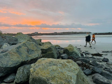 Two people walking along a sandy beach at sunrise during an Ali Knight retreat, seen from a distance beside large coastal rocks, with a vivid pink and orange sky reflected on the wet sand and a bay stretching out behind them