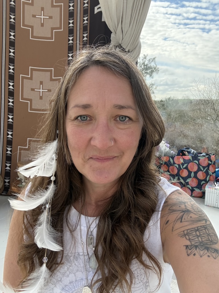 Ali Knight, a white woman with long hair and a nose ring, looking directly at the camera, wearing white feathers in her hair, a white top and layered silver necklaces, with a geometric tattoo visible on her upper arm and a woven geometric wall hanging and rugged Welsh landscape visible behind her