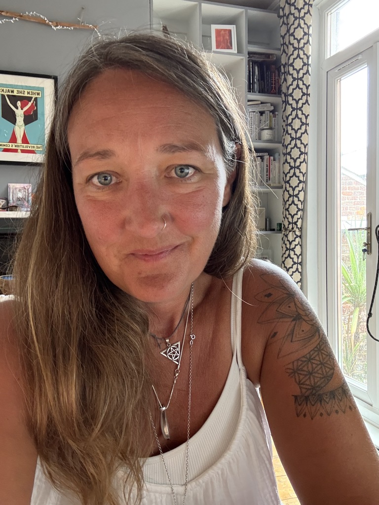 Ali Knight, a white woman with long hair and a nose ring, photographed at home, looking directly at the camera, in a white vest top and layered silver necklaces, with a geometric tattoo visible on her upper arm, bookshelves and a feminist poster reading "When She Walks, The Revolution's Coming" visible in the background