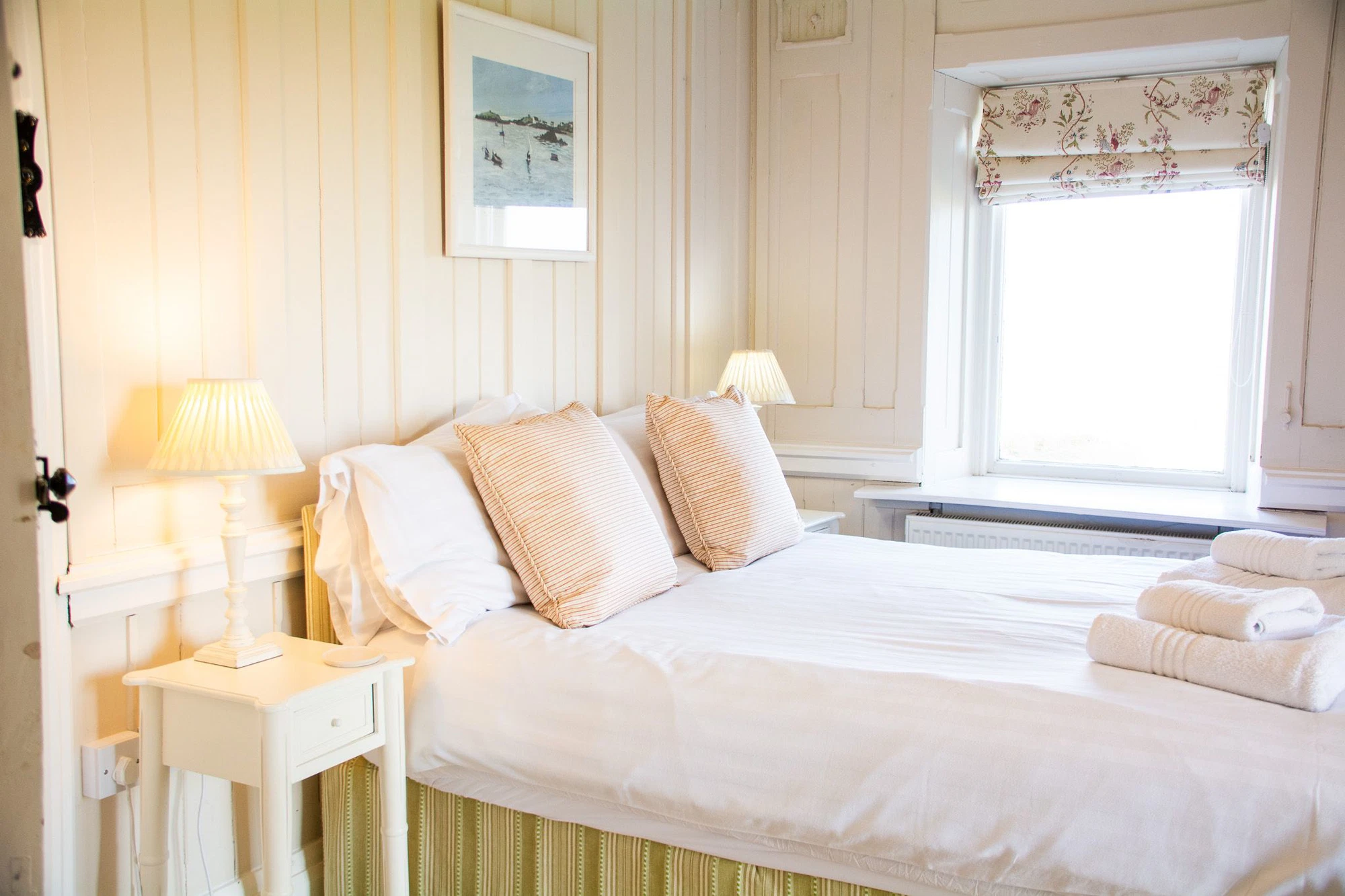 A bright, simply furnished bedroom at Plas Rhoscolyn, Ali Knight's retreat venue, with a double bed dressed in crisp white linen and striped cushions, bedside lamps, folded white towels and a floral roman blind at the window