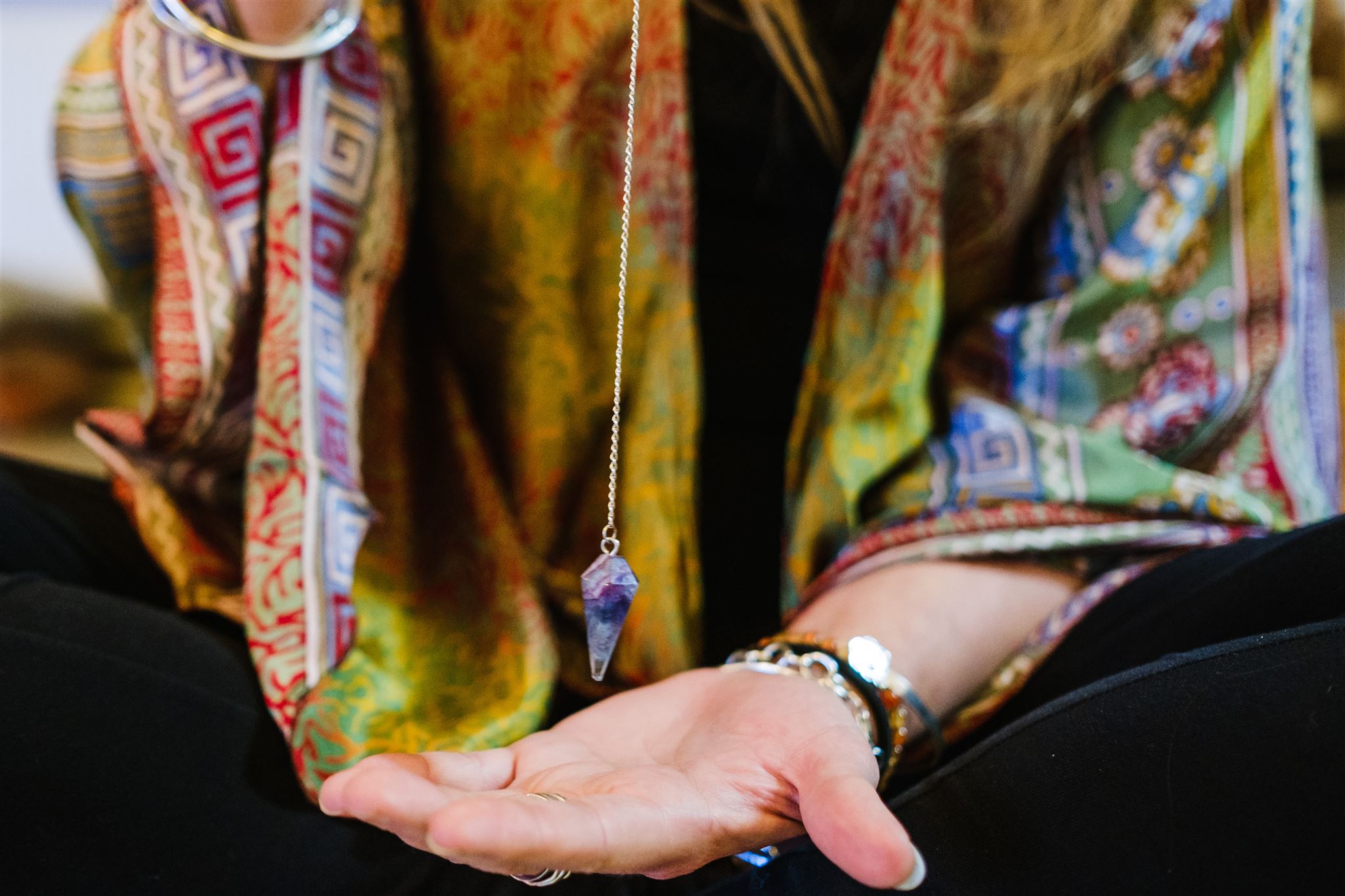 A close-up of Ali Knight's open hand holding a crystal pendulum on a fine silver chain, wearing beaded bracelets and a ring, with her patterned kimono visible in the background