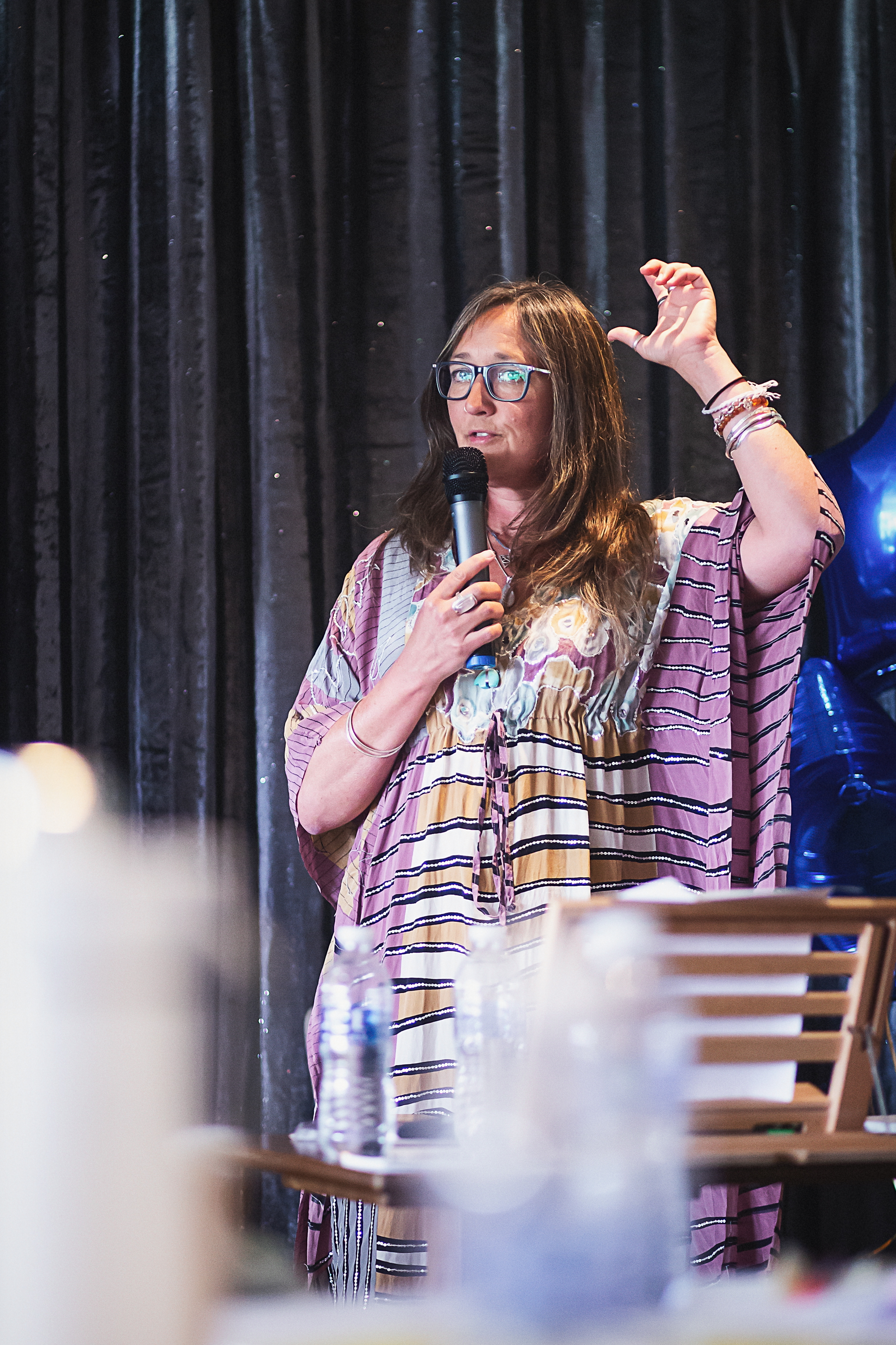 Ali Knight, a white woman with long hair and glasses, speaking into a microphone on stage at a keynote event, wearing a patterned kimono top, one hand raised expressively, with a dark curtain backdrop and a blue balloon visible behind her