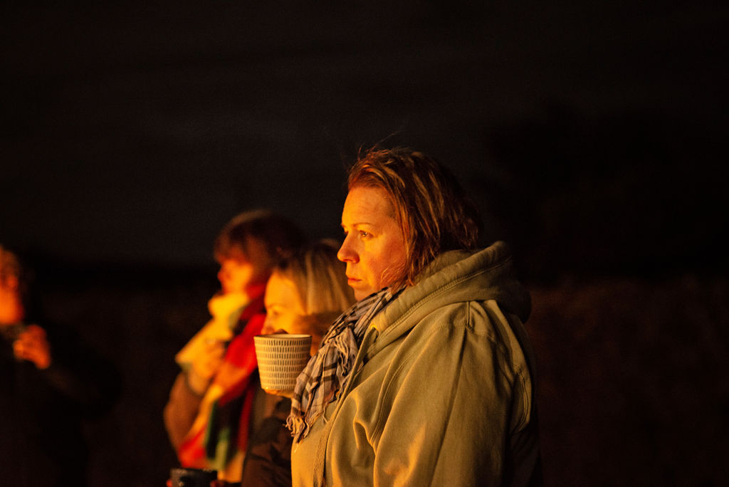 A white woman with windswept hair, holding a steaming mug and gazing into the distance with a calm, reflective expression, lit by warm firelight during an Ali Knight retreat evening, two other people visible in the background