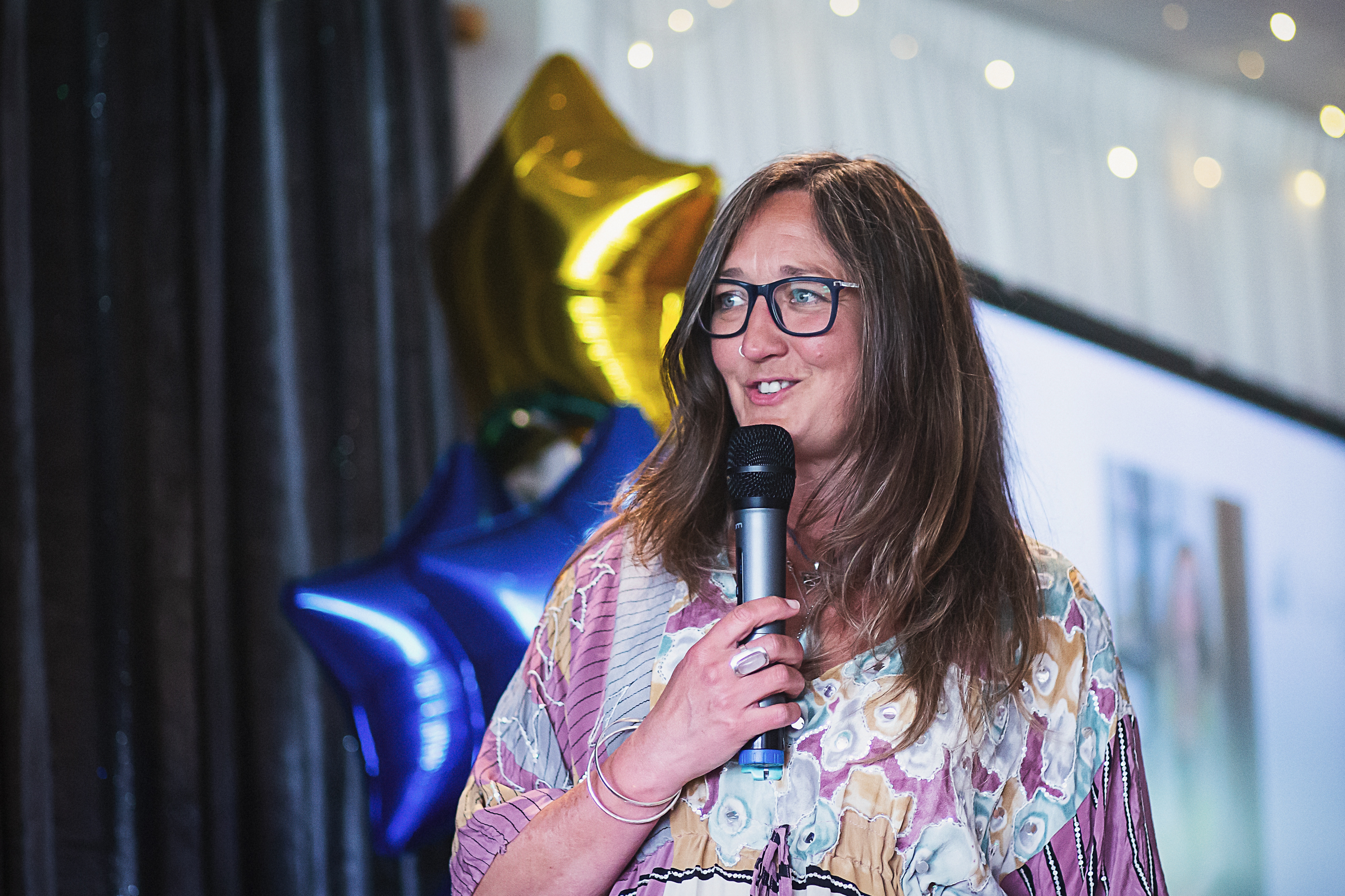Ali Knight, a white woman with long hair and glasses, speaking into a microphone at an event, wearing a patterned kimono top, with gold and blue star-shaped balloons and fairy lights visible behind her