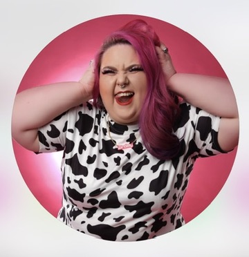 A white female-presenting person with long pink hair, laughing with hands raised to their head in an expressive, joyful pose, wearing a black and white cow print top, photographed against a pink background in a circular crop