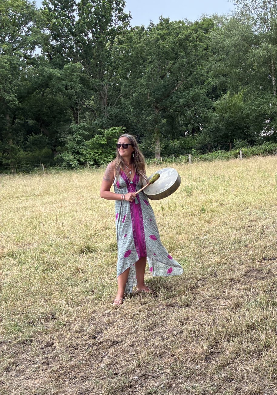 Ali Knight, a white woman with long blonde flecked hair, standing barefoot in a sun-dried meadow playing a shamanic drum, wearing sunglasses, a flowing patterned maxi dress and layered silver necklaces, with a geometric tattoo visible on her upper arm and trees in the background