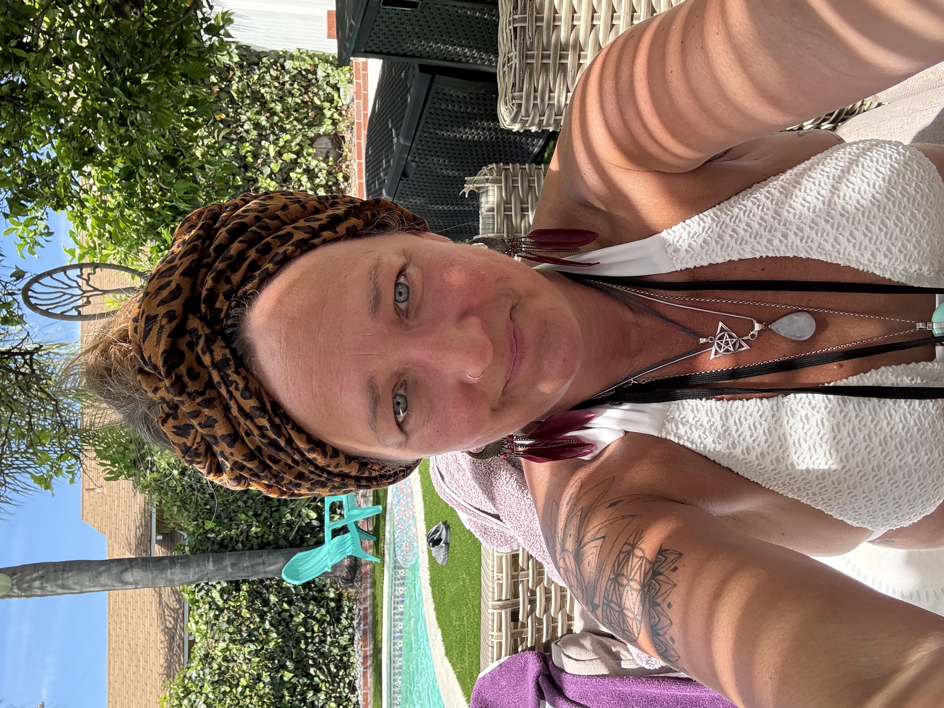 Ali Knight, a white woman with a nose ring, looking directly at the camera, wearing a leopard print head wrap, long red feather earrings, a white top and layered silver necklaces, with a geometric tattoo visible on her upper arm, seated outdoors beside a swimming pool in bright sunshine
