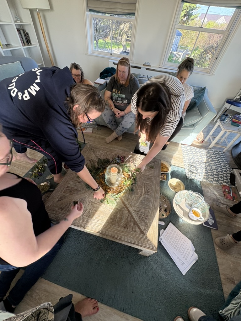 A group of people of mixed ethnicities gathered around a coffee table during an Ali Knight retreat, arranging foliage, dried flowers and a candle into a nature-based centrepiece, with others sitting cross-legged on the floor in a bright, relaxed living room space behind them
