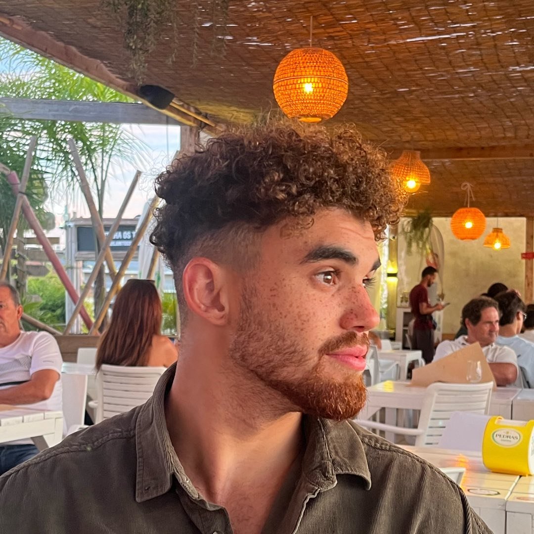 A mixed heritage man with curly hair and a beard, looking to one side with a thoughtful expression, wearing an olive shirt, seated in a warmly lit open-air restaurant with wicker pendant lights and other diners visible in the background