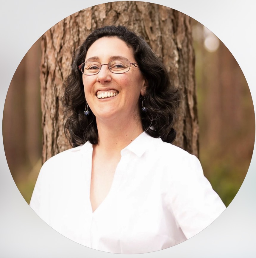 A white woman with short dark curly hair and glasses, smiling broadly while leaning against a tree trunk outdoors, wearing a white shirt and drop earrings, photographed in a circular crop