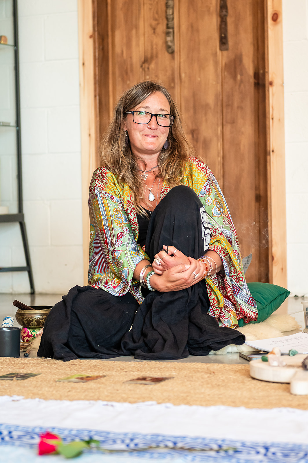 Ali Knight sitting on the floor with knees drawn up, hands clasped, wearing a patterned kimono and silver jewellery, gazing upward with a calm, reflective expression. A singing bowl, crystals, a rose and ceremonial objects are arranged around her.