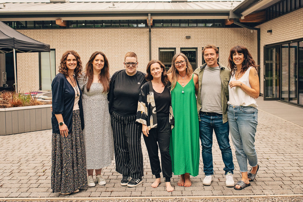 Ali Knight, a white woman in a bright green maxi dress, standing barefoot with a group of six white people of mixed genders outside a modern building at one of her events, all smiling warmly at the camera