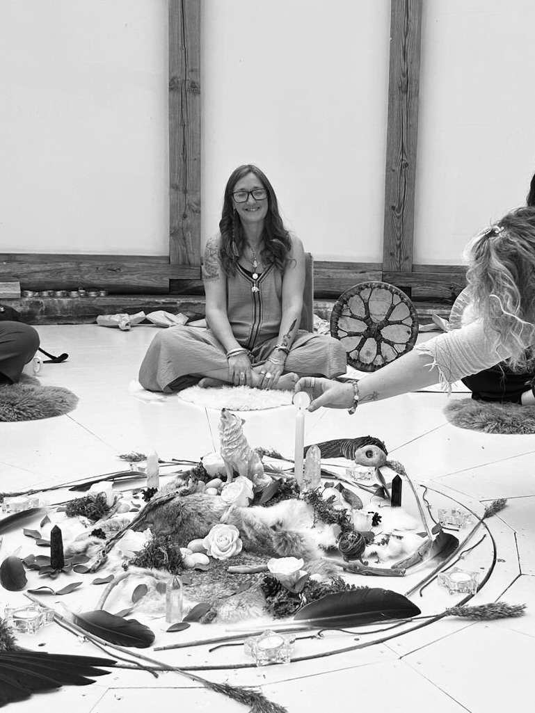 Ali Knight, a white woman with long hair and glasses, sitting cross-legged and smiling during a ceremony, photographed in black and white. An elaborate altar in the foreground contains crystals, feathers, flowers, candles, animal figures and natural objects arranged in a circle. Other participants are partially visible around the edges of the space, with a shamanic drum visible to one side