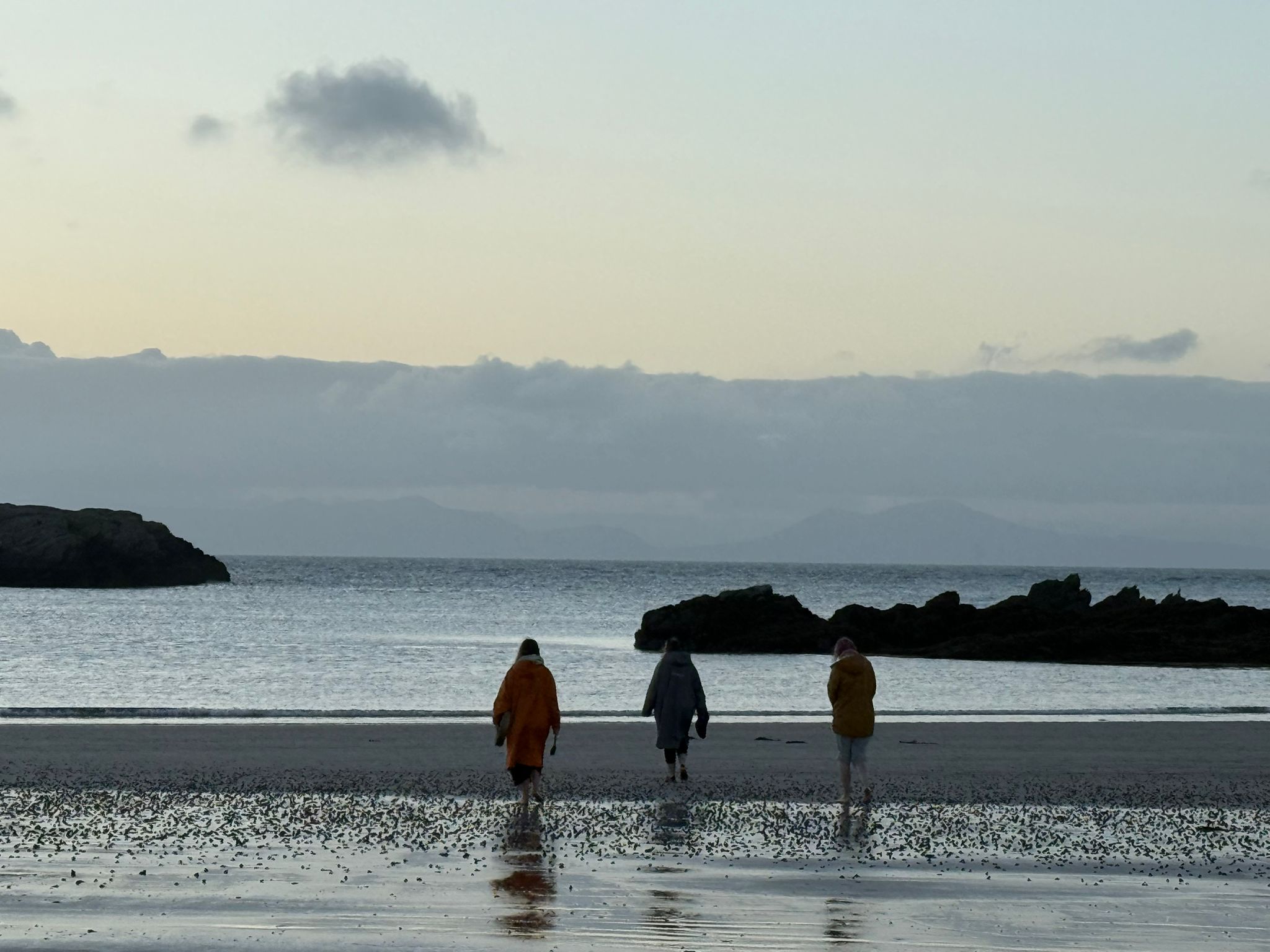 Three people walking along a deserted beach at dawn during an Ali Knight retreat, seen from behind, wearing brightly coloured coats, with rocky outcrops, a calm sea and mountains faintly visible on the horizon in the early morning light