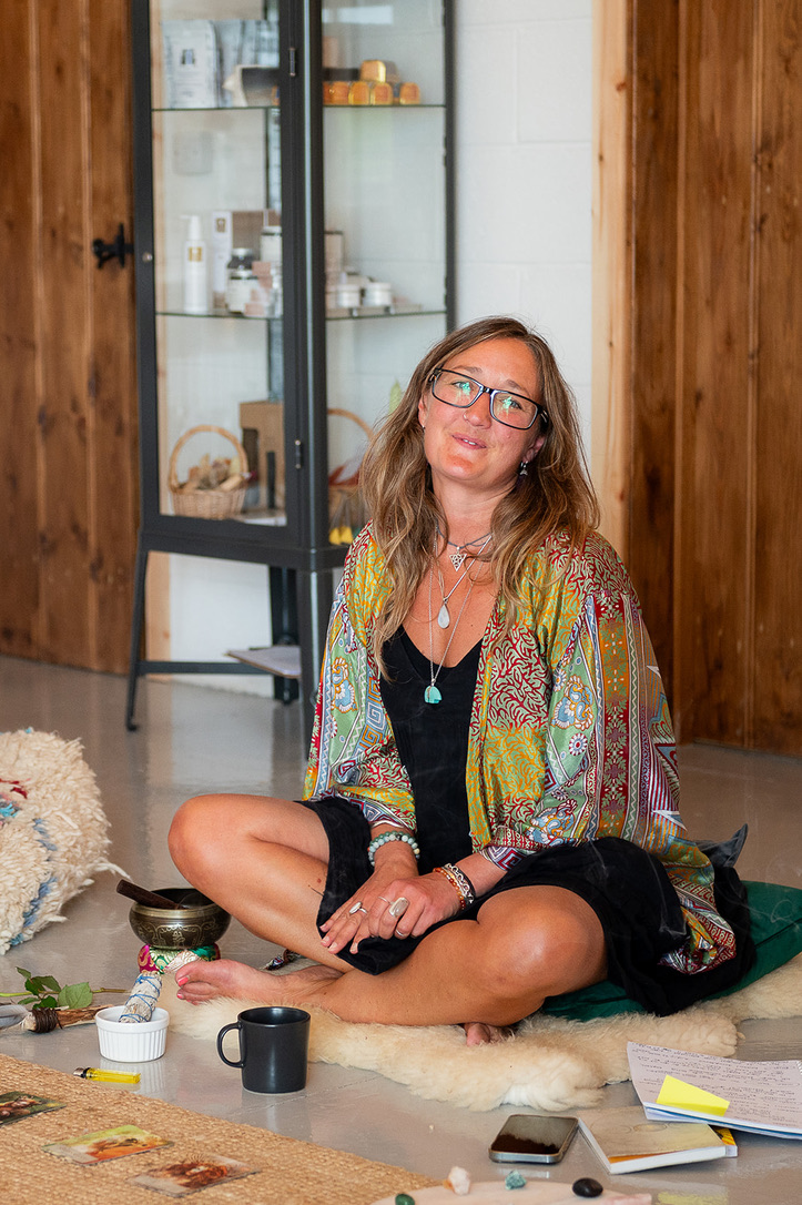 Ali Knight, soul coach, sitting cross-legged on a sheepskin rug surrounded by journals, crystals, a singing bowl and a mug, wearing a patterned kimono and layered silver necklaces, looking directly at the camera with a relaxed, open expression.