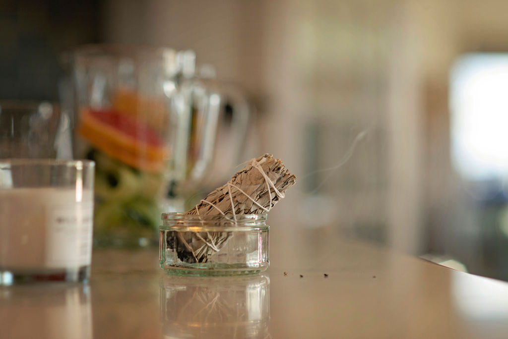 A white sage smudge stick resting in a small glass jar on a surface, with a lit candle and a glass bowl of flowers softly out of focus in the background, a thin wisp of smoke rising from the sage