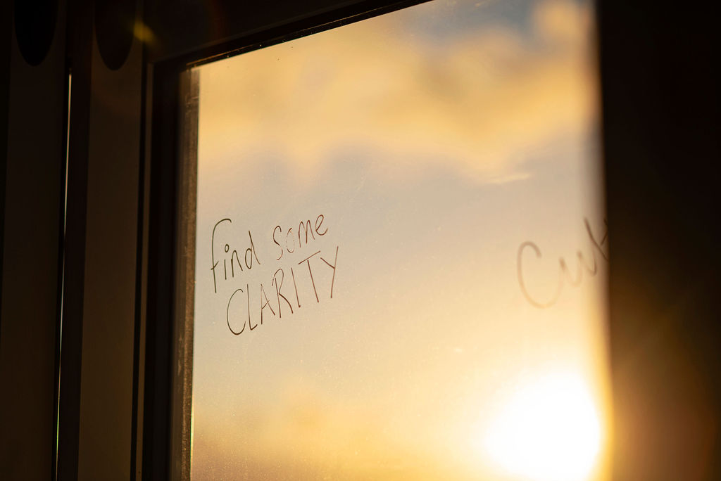 The words "find some CLARITY" written in chalk pen on a window, with warm golden sunrise light and clouds visible through the glass