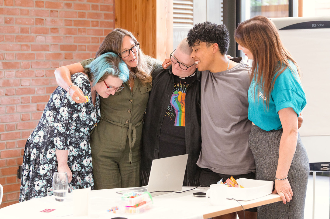 Five people standing together with arms around each other, laughing and smiling during an Ali Knight group event in a bright indoor space with exposed brick walls. The group includes white people and a mixed-race person, with a range of styles including a rainbow pride t-shirt and blue-tipped hair, reflecting a warm and diverse gathering.