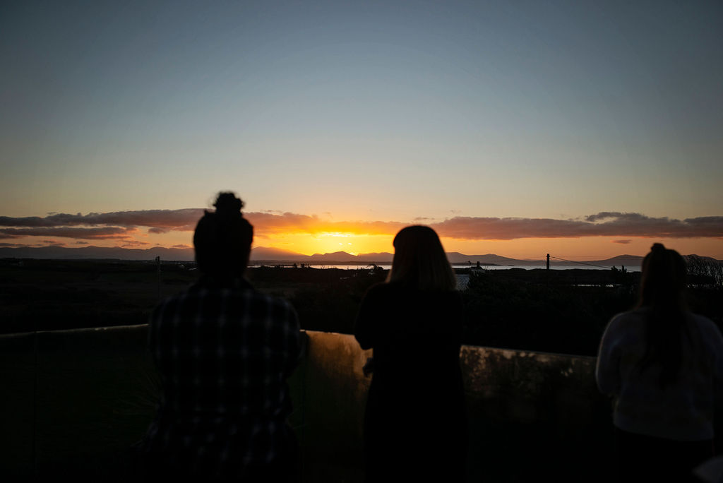 Three people silhouetted against a dramatic sunset over the sea and mountains during an Ali Knight retreat, the sky glowing orange and gold on the horizon with soft clouds above