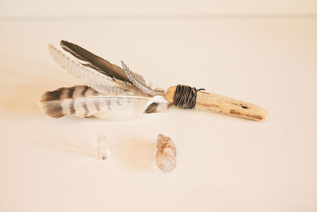 A handmade feather wand with striped grey and white feathers bound to a piece of driftwood with dark cord, resting alongside a small raw crystal on a pale surface