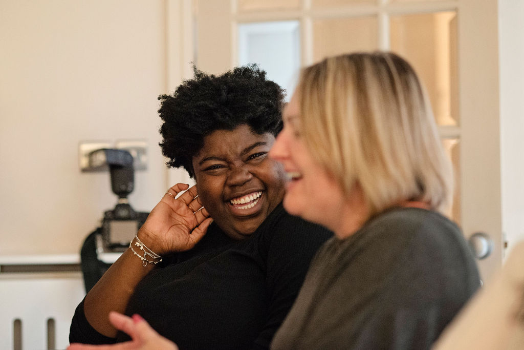 A Black woman with natural hair laughing joyfully with her hand raised to her face, alongside a white woman with blonde hair who is also laughing, both sharing a warm spontaneous moment during an Ali Knight group event