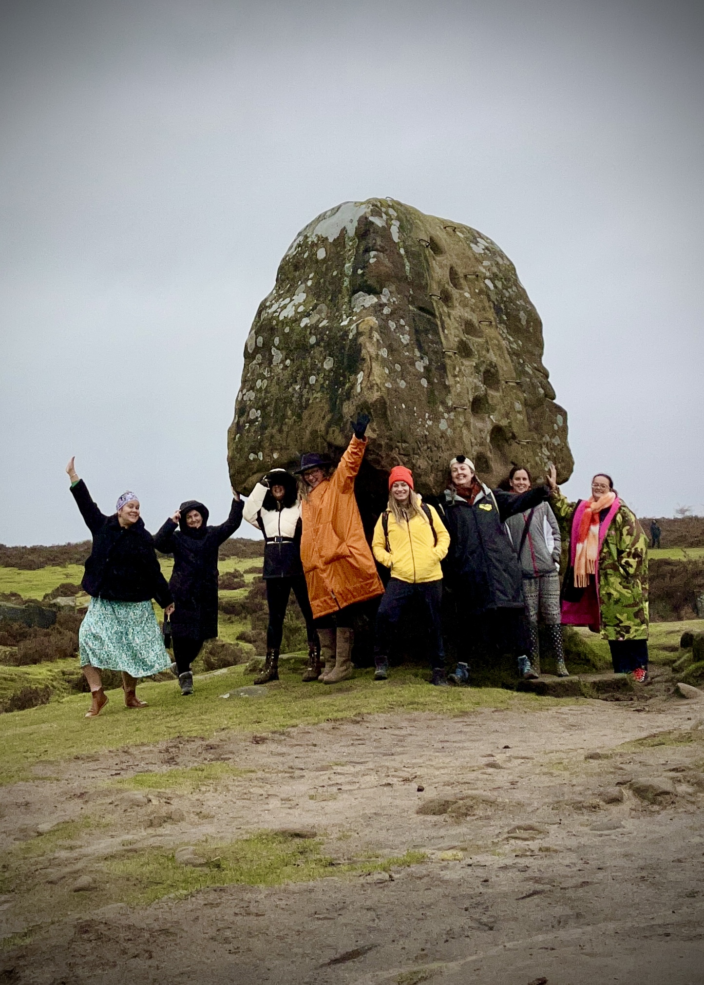 A group of people of mixed ethnicities gathered around a large ancient standing stone on the moorland near Nine Ladies Stone Circle in the Peak District, dressed in colourful winter coats and hats, laughing and raising their arms in celebration during an Ali Knight retreat