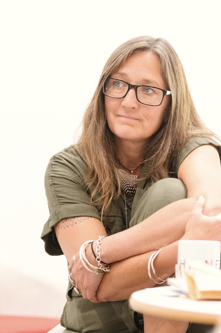 Ali Knight, a white woman with long hair and glasses, sitting with arms folded and resting on her knees, wearing an olive green top, silver bracelets and a feather pendant necklace, gazing thoughtfully into middle distance with a calm, present expression.