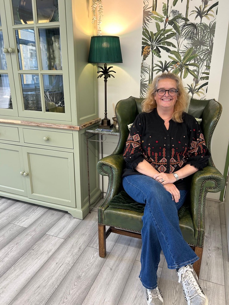 A white woman with blonde hair and glasses, sitting in a green leather wingback chair, smiling warmly at the camera, wearing a black embroidered top, jeans and trainers, with a green dresser, palm print wallpaper and a table lamp visible behind her