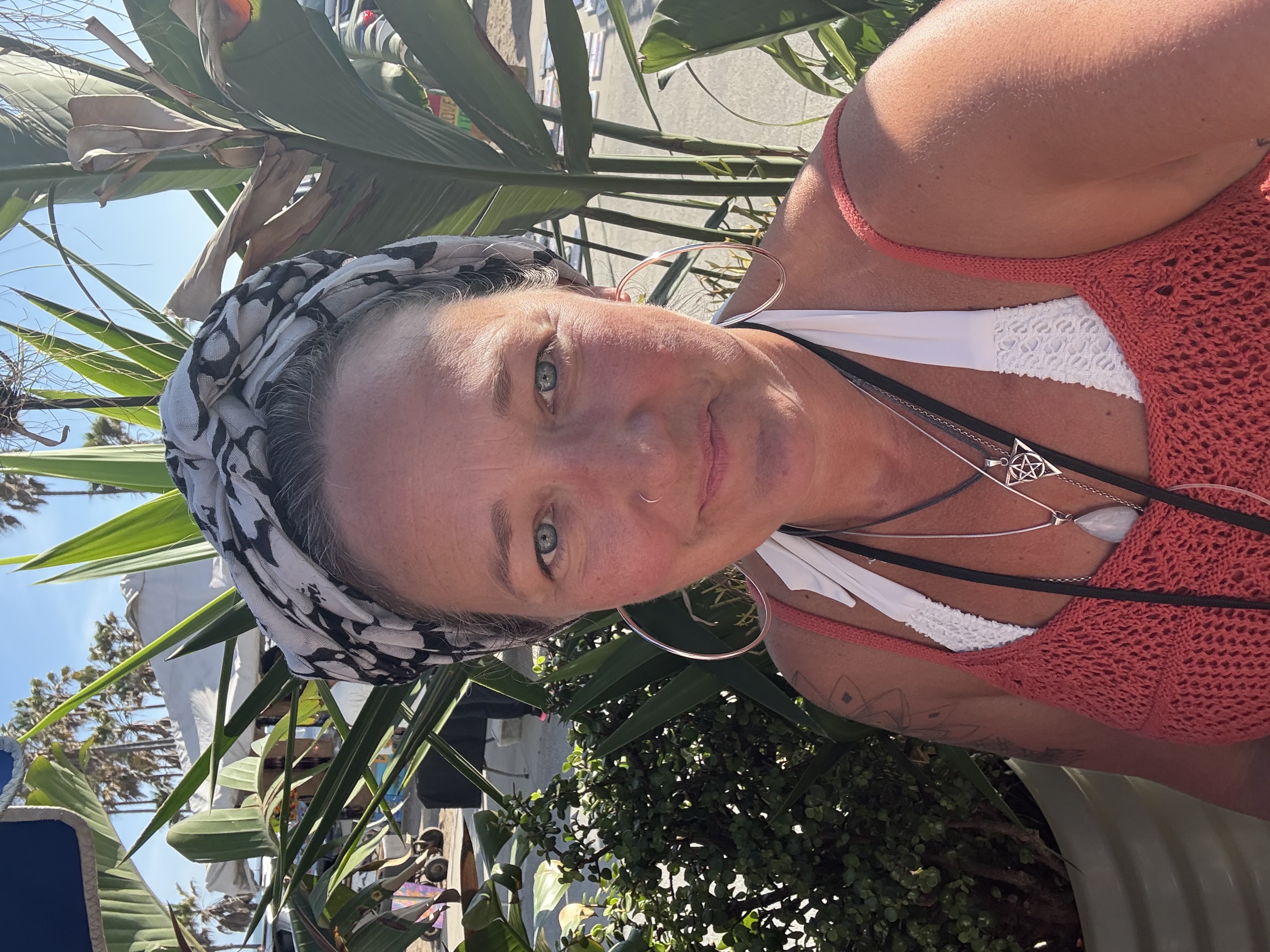 Ali Knight outdoors in bright sunshine, wearing a patterned head wrap, large hoop earrings and layered silver necklaces, looking at the camera and smiling, with tropical plants and a sunny outdoor space visible behind her.