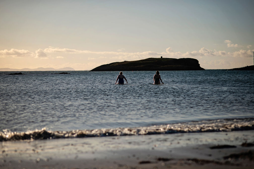 Two people wading into the sea from a sandy beach during an Ali Knight retreat, seen from behind in silhouette, with a small island, mountains and a wide open sky visible on the horizon