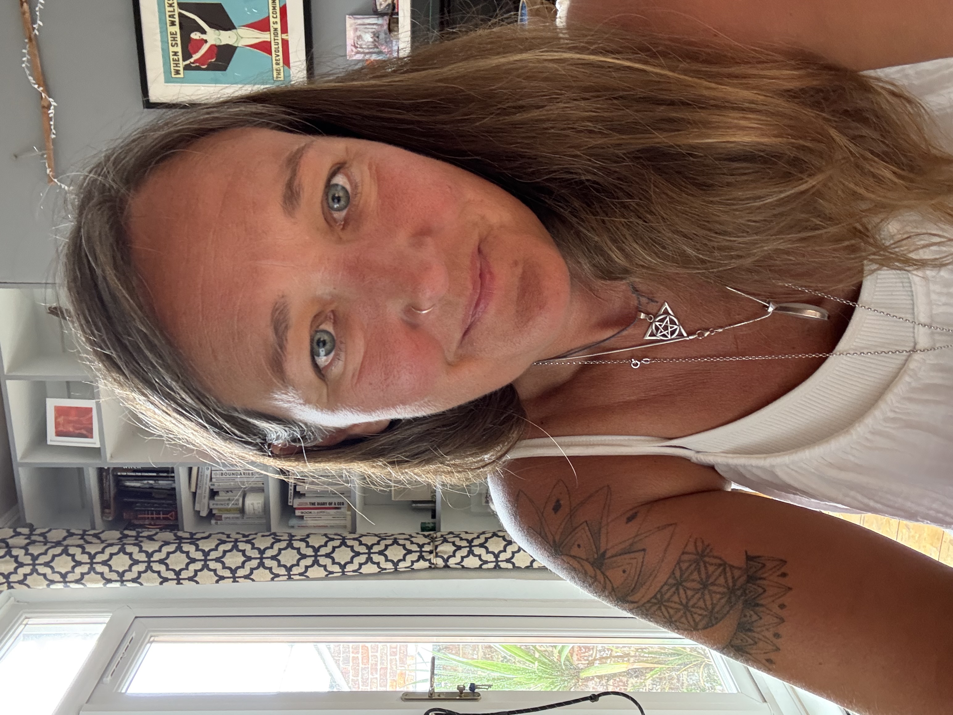 Ali Knight, a white woman with long hair and a nose ring, photographed at home, looking directly at the camera, in a white vest top and layered silver necklaces, with a geometric tattoo visible on her upper arm, bookshelves and a feminist poster reading "When She Walks, The Revolution's Coming" visible in the background