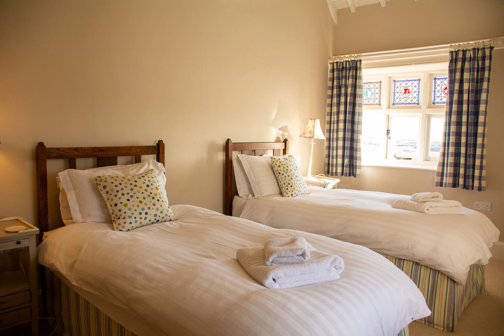 A warm, characterful twin bedroom at Plas Rhoscolyn, Ali Knight's retreat venue, with two single beds dressed in white linen and spotty cushions, wooden headboards, folded towels, a bedside lamp and a window with stained glass panels and checked curtains
