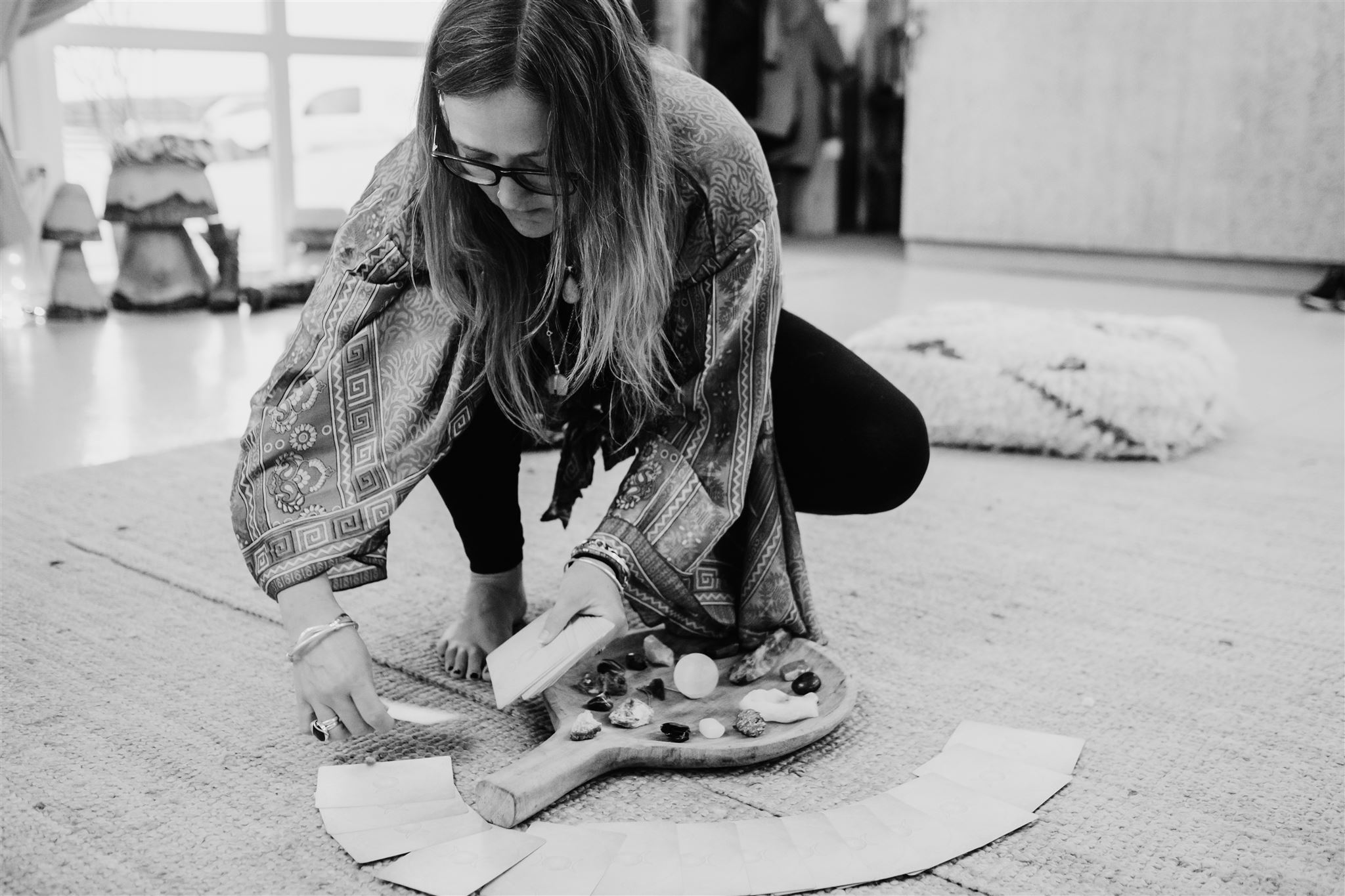 Ali Knight, a white woman with long hair and glasses, crouching on the floor to arrange crystals, stones and a piece of selenite on a wooden board, photographed in black and white, with cushions and a light-filled space visible in the background