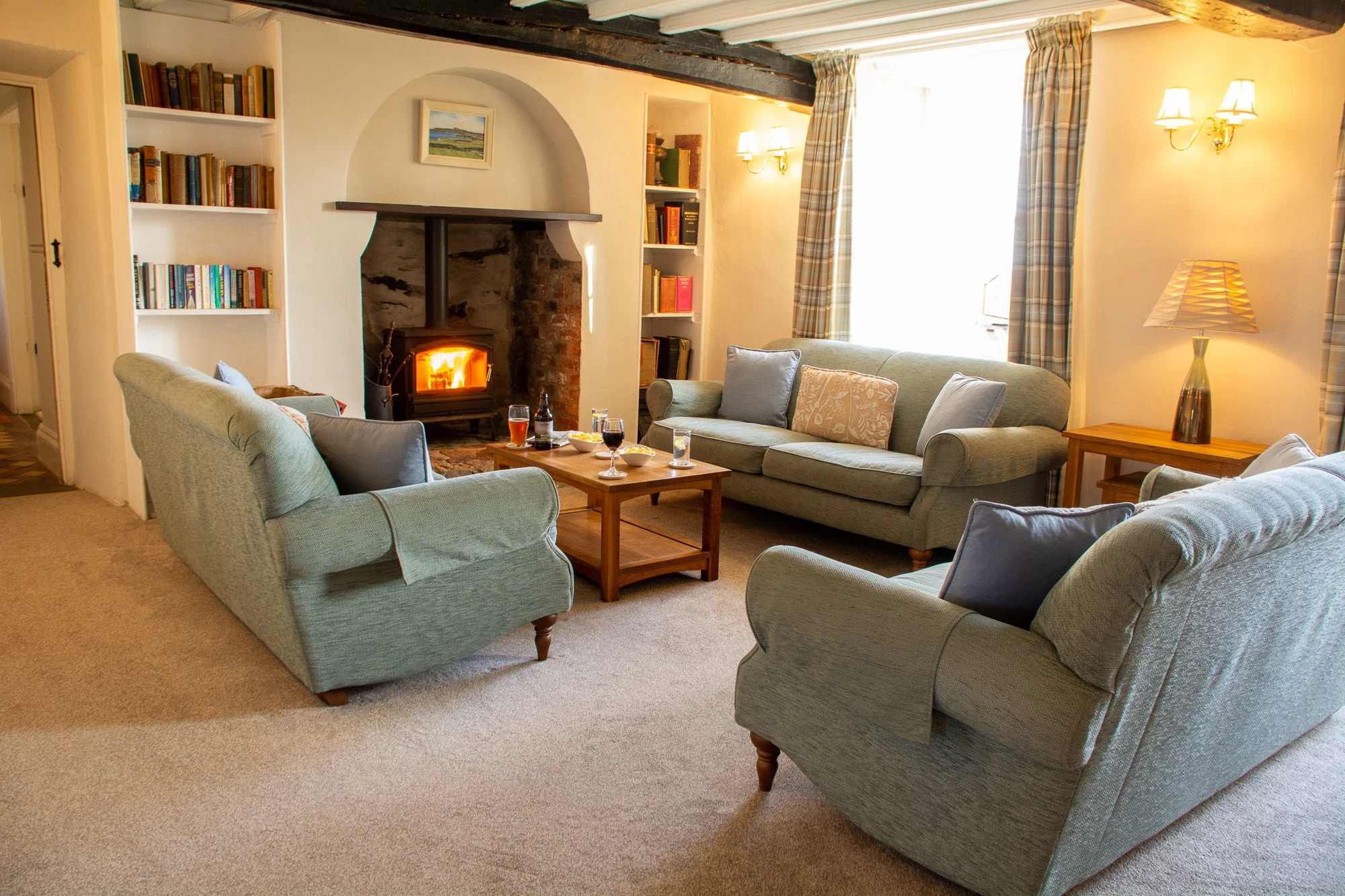 The cosy sitting room at Plas Rhoscolyn, Ali Knight's retreat venue, with soft grey sofas, a glowing wood burning stove set into an original stone fireplace, bookshelves, warm lamplight and drinks on the coffee table