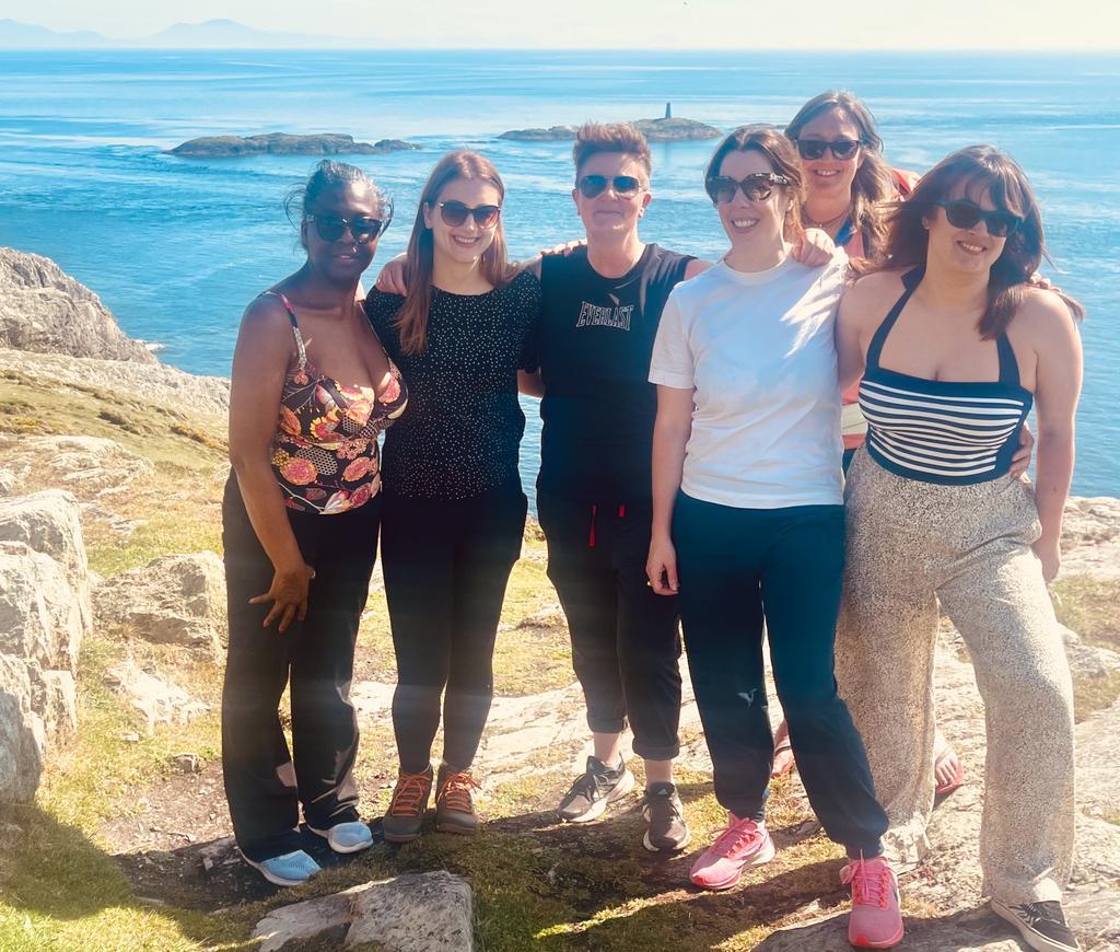 A diverse group of six people of mixed ethnicities standing together on coastal rocks during an Ali Knight retreat, smiling and wearing sunglasses, with a bright blue sea, rocky outcrops and a distant lighthouse visible behind them on a sunny day