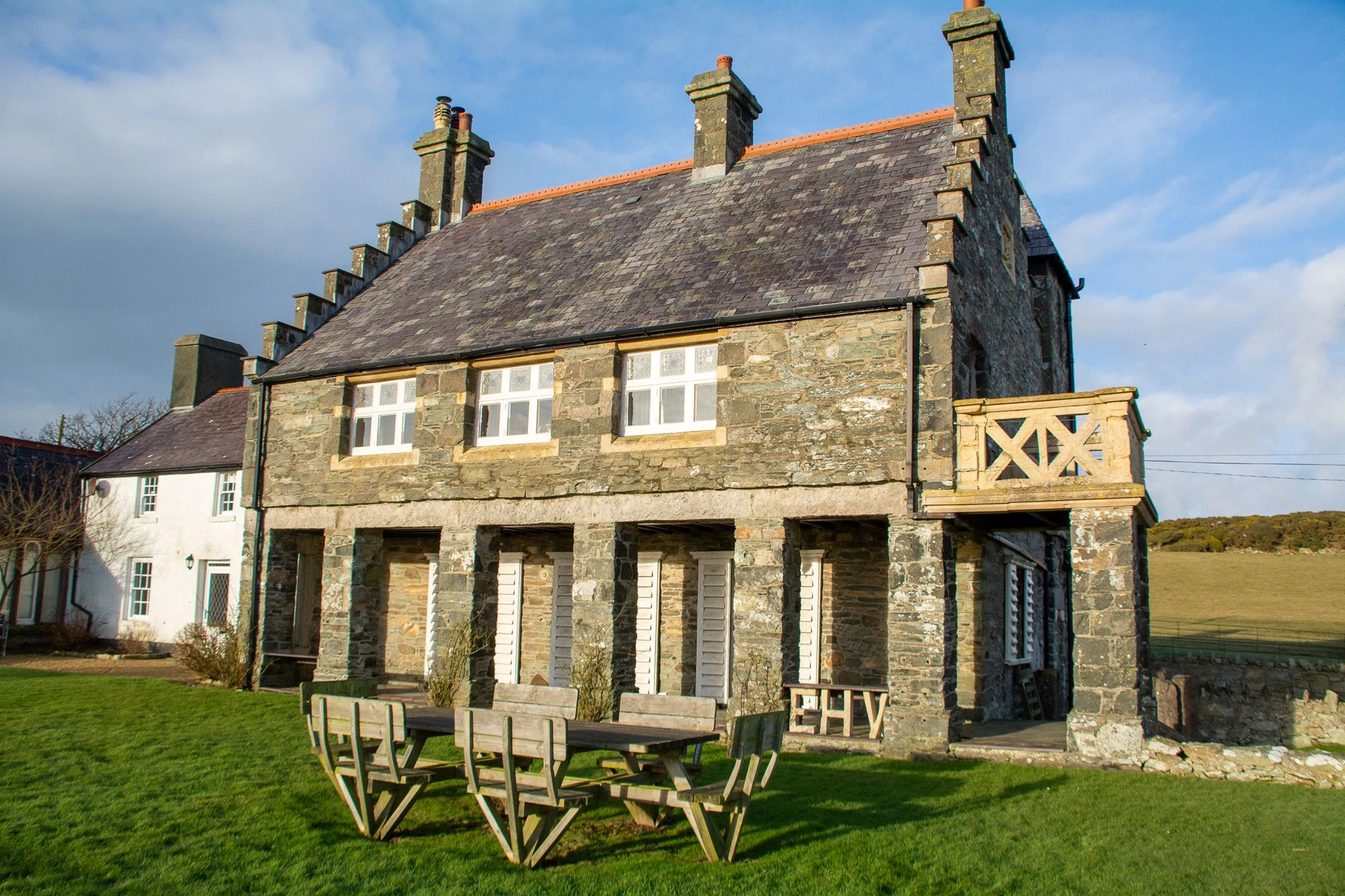 The exterior of Plas Rhoscolyn, Ali Knight's retreat venue in Wales, a historic stone building with a slate roof, white-shuttered windows and a wooden balcony, with picnic benches on the lawn and open countryside visible behind under a bright blue sky