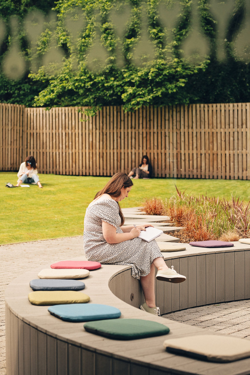 A white woman with long brown hair sitting alone on a curved outdoor bench, writing in a journal during an Ali Knight event, wearing a spotted dress, with two other white women sitting separately on the lawn in the background, surrounded by lush greenery and colourful seat cushions