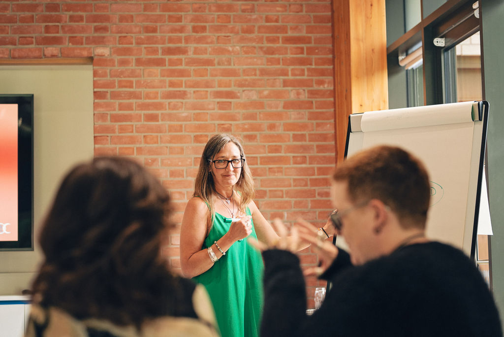 Ali Knight, a white woman with long hair and glasses, facilitating a group session in a bright room with exposed brick walls, wearing a green dress and silver jewellery, with a flipchart beside her and participants visible in the foreground