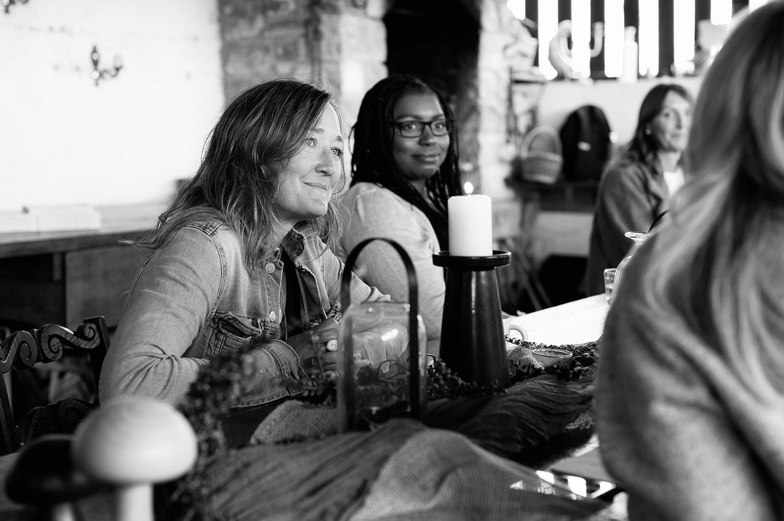 Ali Knight, a white woman with long hair, gazing attentively forward during a group event, photographed in black and white. A Black woman with locs and glasses smiles behind her, with candles and natural objects on the table and other participants visible in the background