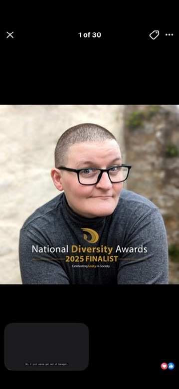 A white non-binary person with a shaved head and glasses, smiling warmly at the camera, with a National Diversity Awards 2025 Finalist graphic overlaid on the image