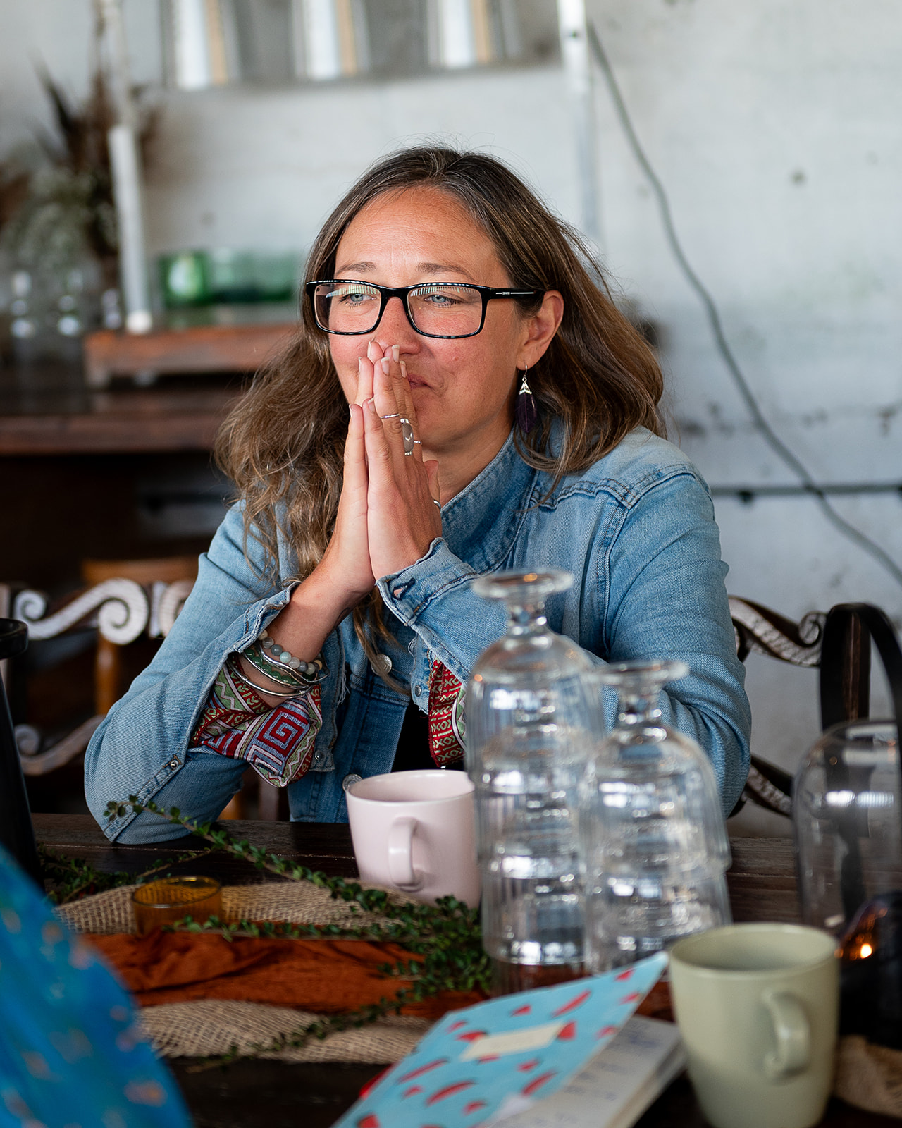 Ali Knight, a white woman with long hair and glasses, sitting at a table with her hands pressed together in a thoughtful, listening pose, wearing a denim jacket over a patterned top and feather earrings, with glasses, mugs, water jugs and foliage on the table in front of her
